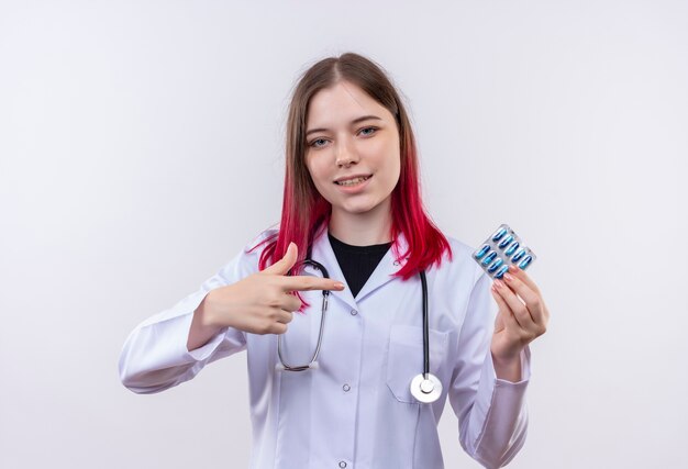 Felice giovane medico ragazza indossa stetoscopio abito medico punta a pillole in mano su sfondo bianco isolato
