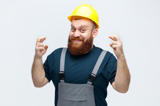 Felice giovane operaio edile maschio che indossa casco di sicurezza e uniforme guardando la fotocamera mentre incrocia le dita isolate su sfondo bianco