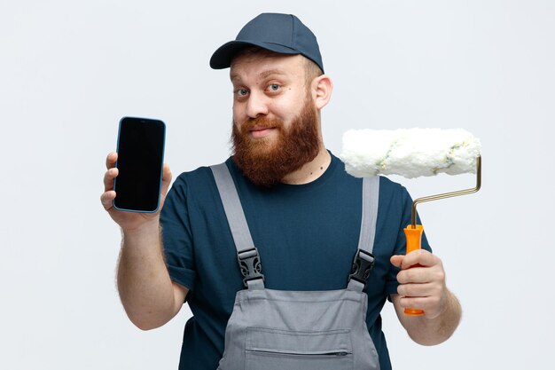 Felice giovane operaio edile maschio che indossa il cappuccio e uniforme che tiene il rullo di vernice guardando la fotocamera che mostra il telefono cellulare alla fotocamera isolata su sfondo bianco