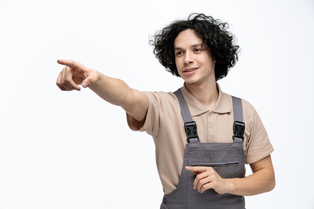 Felice giovane operaio edile maschio che indossa l'uniforme guardando il lato che punta le dita a lato isolato su sfondo bianco