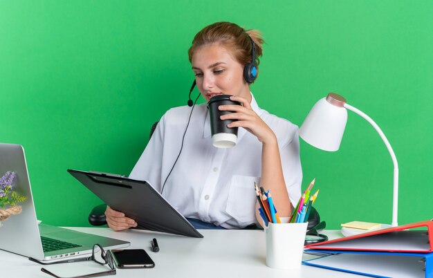 Felice giovane ragazza bionda del call center che indossa l'auricolare seduto alla scrivania con strumenti di lavoro che tengono tazza di caffè in plastica e appunti guardando appunti isolati sul muro verde