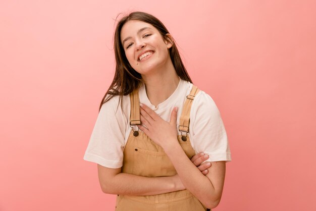 Felice giovane ragazza bruna caucasica che tiene la mano sul petto sorridendo alla telecamera su sfondo rosa Concetto di emozioni e stati d'animo