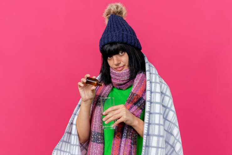 Foto gratuito felice giovane ragazza caucasica malata indossando cappello invernale e sciarpa avvolto in plaid versando medicamento in vetro in un bicchiere d'acqua guardando la telecamera isolata su sfondo cremisi con spazio di copia