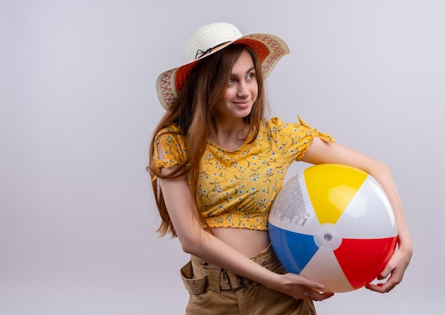 Felice giovane ragazza che indossa un cappello tenendo palla spiaggia guardando il lato destro su uno spazio bianco isolato con spazio di copia