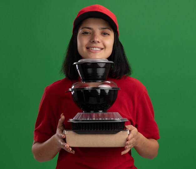 Felice giovane ragazza delle consegne in uniforme rossa e cappuccio che tiene pila di confezioni di cibo sorridente amichevole in piedi sopra la parete verde