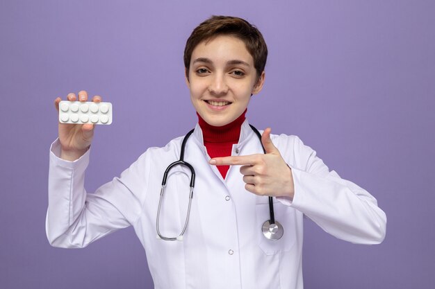 Felice giovane ragazza dottore in camice bianco con stetoscopio intorno al collo tenendo blister con pillole puntate con il dito indice su di esso sorridente fiducioso in piedi sul viola