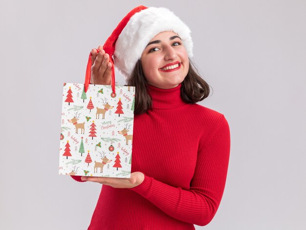 Felice giovane ragazza in maglione rosso e cappello santa tenendo il sacchetto di carta colorato con i regali di Natale guardando la fotocamera con il sorriso sul viso in piedi su sfondo bianco