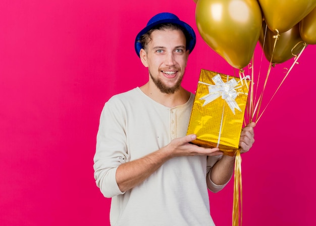 Felice giovane ragazzo slavo bello del partito che indossa il cappello del partito che tiene i palloncini e mostrando il contenitore di regalo che esamina la parte anteriore isolata sulla parete rosa con lo spazio della copia