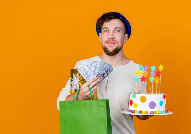 Felice giovane ragazzo slavo bello partito che indossa il cappello del partito che tiene il sacchetto di carta dei soldi del contenitore di regalo e torta di compleanno con le stelle che guarda l'obbiettivo isolato su fondo arancio con lo spazio della copia