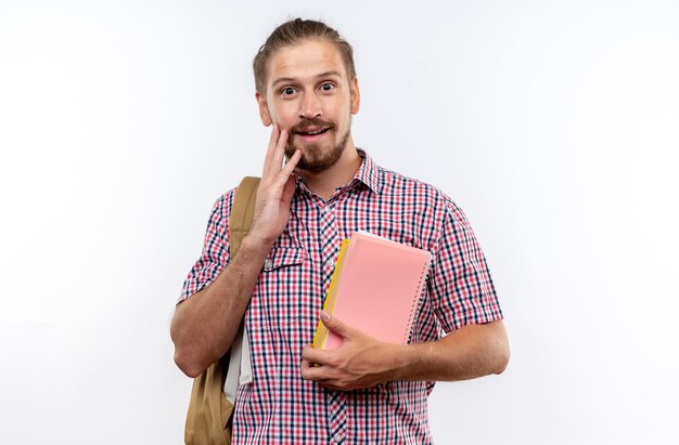 Felice giovane studente che indossa uno zaino con in mano libri che mettono la mano sulla guancia