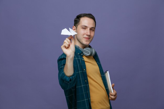 Felice giovane studente maschio che indossa le cuffie intorno al collo in piedi nella vista di profilo tenendo il blocco note guardando la fotocamera che mostra l'aeroplano di carta isolato su sfondo viola