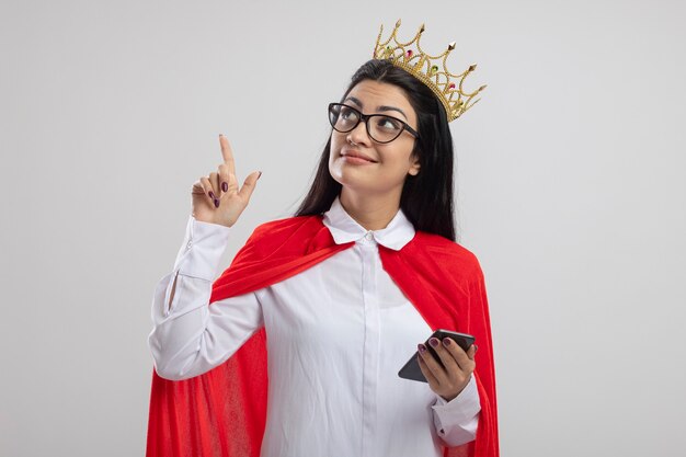 Felice giovane superdonna con gli occhiali e corona che tiene il telefono cellulare alla ricerca e rivolto verso l'alto isolato sul muro bianco