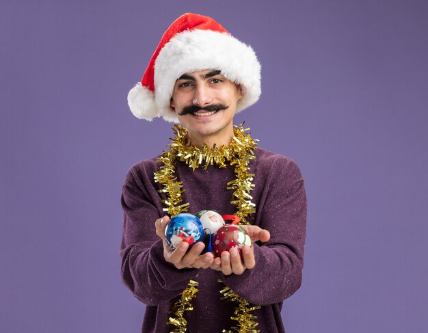 Felice giovane uomo baffuto che indossa il cappello di Babbo Natale con orpelli intorno al collo tenendo le palle di Natale felice e allegro sorridente in piedi sul muro viola