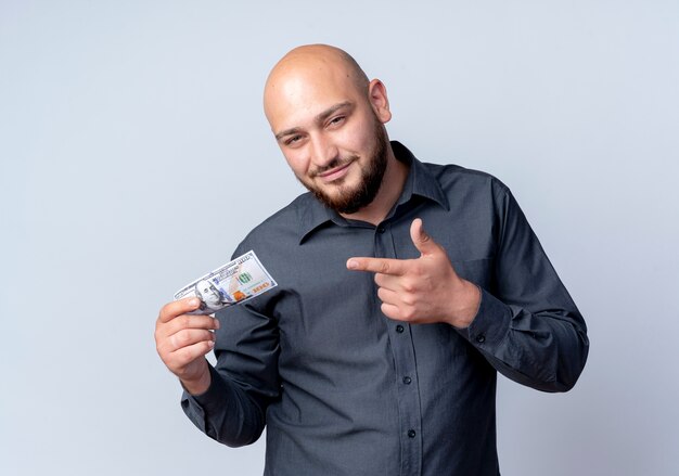 Felice giovane uomo calvo call center holding e indicando denaro isolato su sfondo bianco