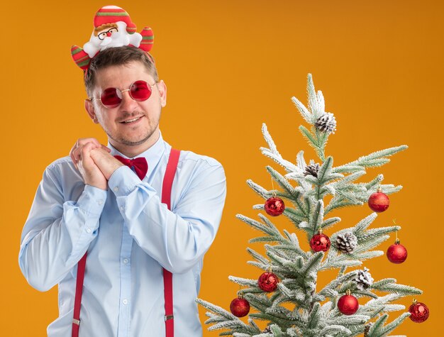 Felice giovane uomo che indossa bretelle farfallino nel cerchio con Babbo Natale e occhiali rossi in piedi accanto all'albero di natale tenendo le mani insieme in attesa di sorpresa su sfondo arancione