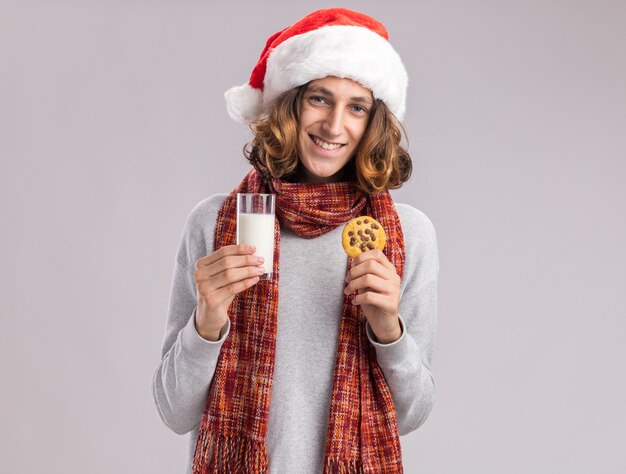 Felice giovane uomo che indossa il cappello di Babbo Natale con sciarpa calda intorno al collo tenendo un bicchiere di latte e biscotto guardando sorridente allegramente