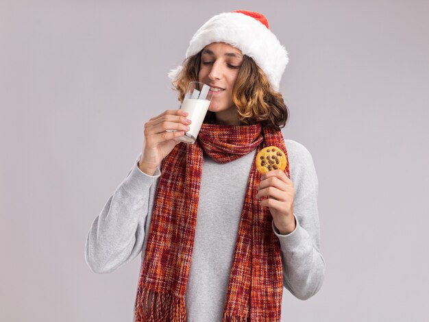 Felice giovane uomo che indossa il cappello di Babbo Natale con una calda sciarpa intorno al collo bere latte in possesso di cookie