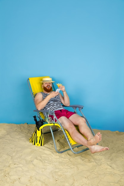 Felice giovane uomo che riposa, prende selfie, bevendo cocktail su sfondo blu studio. Concetto di emozioni umane, espressione facciale, vacanze estive o fine settimana. Freddo, estate, mare, oceano, alcol.