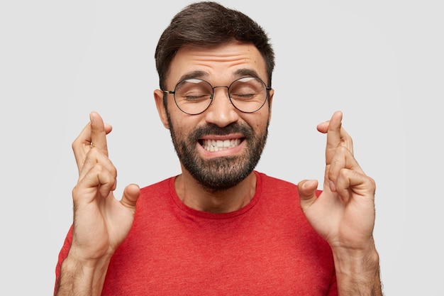 Felice giovane uomo con la barba incolta, incrocia le dita, tiene gli occhi chiusi, vestito con una maglietta rossa