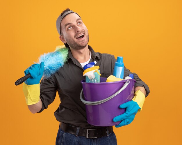 Felice giovane uomo delle pulizie che indossa abiti casual e berretto in guanti di gomma che tiene secchio con strumenti di pulizia e spolverino colorato guardando con il sorriso sul viso in piedi sopra la parete arancione