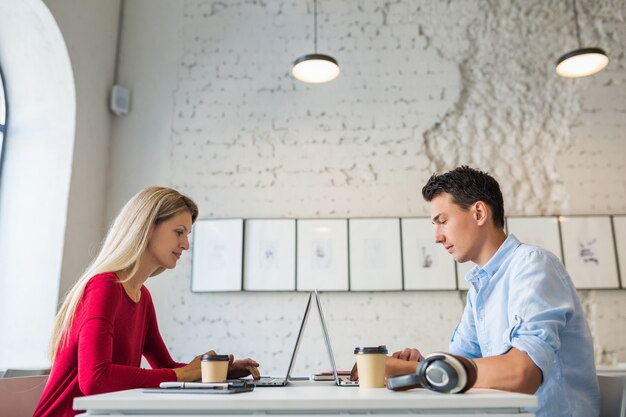 Felice giovane uomo e donna seduta al tavolo faccia a faccia, lavorando al computer portatile in ufficio di co-working