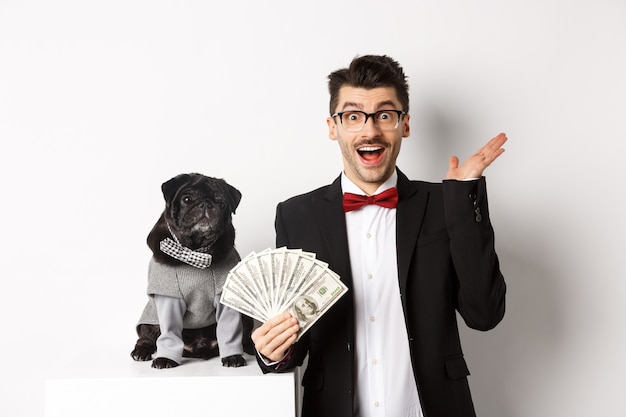 Felice giovane uomo e simpatico cane nero in piedi in costumi da festa, proprietario di un carlino che tiene in mano dollari di denaro, fissando la telecamera stupita, sfondo bianco