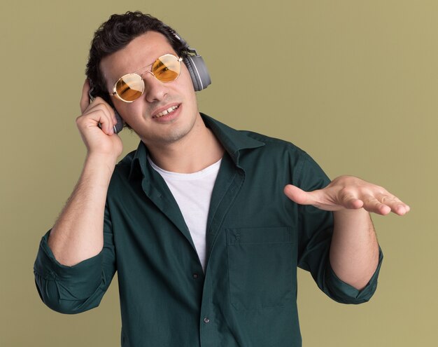 Felice giovane uomo in camicia verde con gli occhiali con le cuffie godendo la sua musica preferita in piedi sopra la parete verde