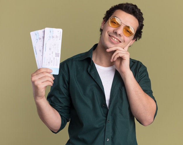 Felice giovane uomo in camicia verde con gli occhiali in possesso di biglietti aerei guardando la parte anteriore con il sorriso sul viso in piedi sopra la parete verde