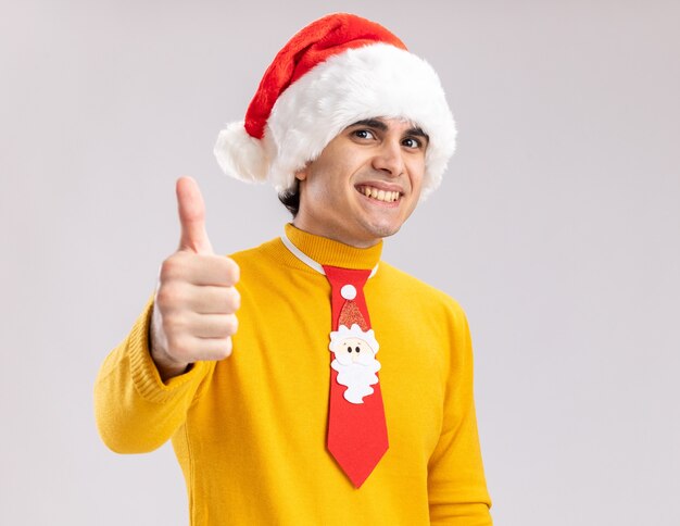 Felice giovane uomo in dolcevita giallo e santa hat con divertente cravatta guardando la telecamera con il sorriso sul viso che mostra i pollici in su in piedi su sfondo bianco