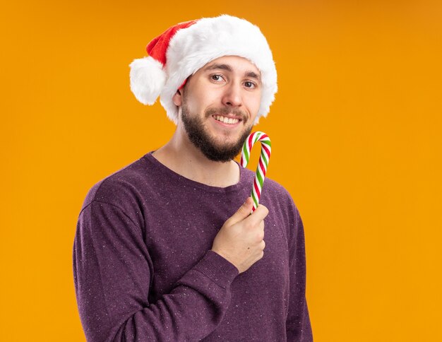 Felice giovane uomo in maglione viola e santa hat tenendo il bastoncino di zucchero guardando la fotocamera con il sorriso sul viso in piedi su sfondo arancione