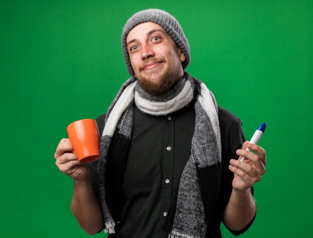 Felice giovane uomo slavo malato con sciarpa intorno al collo indossando cappello invernale con termometro e tazza isolati su parete verde con spazio copia