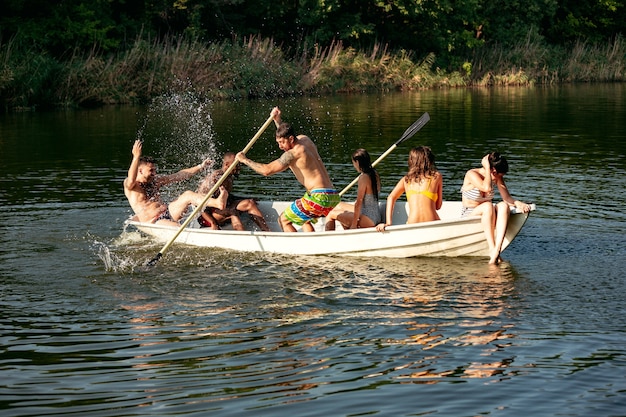 Felice gruppo di amici che si divertono mentre ridono e nuotano nel fiume. Uomini e donne allegri in costume da bagno in una barca in riva al fiume nella giornata di sole. Estate, amicizia, resort, concetto di fine settimana.