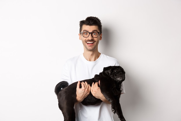 Felice hipster con gli occhiali che tiene carino carlino nero e sorridente, proprietario del cane che fissa la telecamera con un sorriso stupito, in piedi su sfondo bianco.