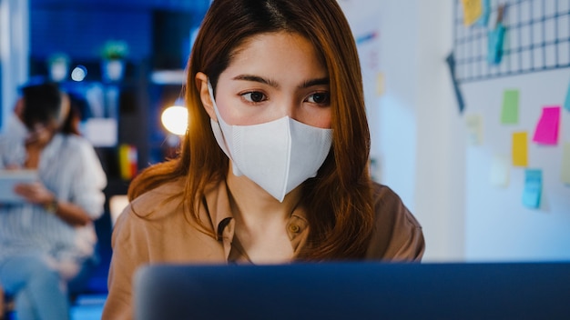 Felice imprenditrice asiatica che indossa una maschera medica per il distanziamento sociale in una nuova situazione normale per la prevenzione dei virus mentre si utilizza il laptop al lavoro durante la notte in ufficio.