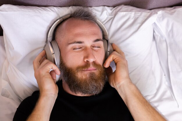 Felice l'uomo caucasico sul letto che indossa la cuffia ascolta goditi la sua musica preferita, riposando da solo, ballando.