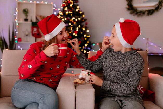 Felice madre e figlio piccolo in cappelli della santa con tazze di tè mangiando biscotti seduti su un divano divertendosi in camera decorata con albero di natale in background