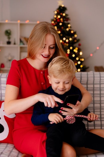 Felice madre in abito rosso con il suo piccolo bambino seduto su un divano con lo smartphone in una stanza decorata con albero di Natale in background