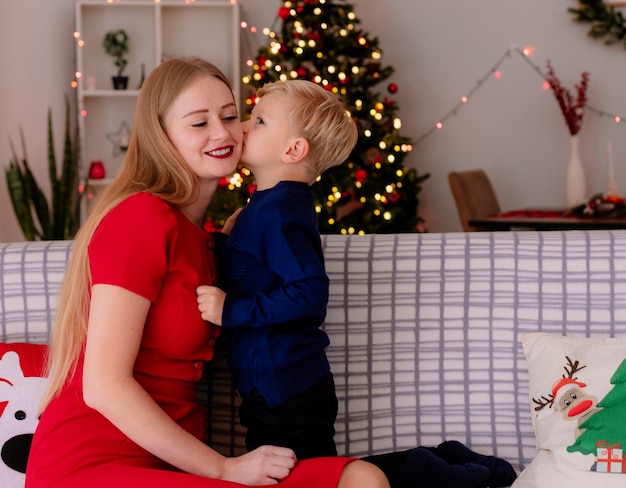 Felice madre in abito rosso con il suo piccolo bambino seduto su un divano piccolo bambino che bacia la sua allegra madre in una stanza decorata con albero di natale in background