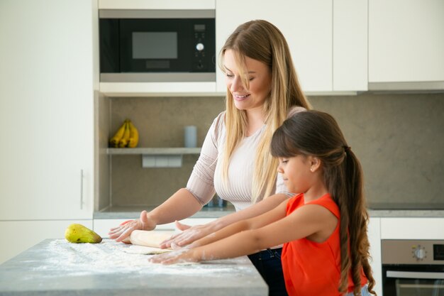 Felice mamma e figlia che rotolano la pasta sul tavolo della cucina. Ragazza e sua madre che cuociono insieme il pane o la torta. Colpo medio, vista laterale. Concetto di cucina familiare