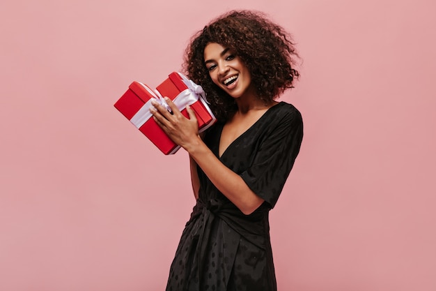 Felice meravigliosa mulatta con i capelli ricci bruna in abito nero a pois sorridente, guardando nella fotocamera e tenendo in mano due scatole regalo rosse