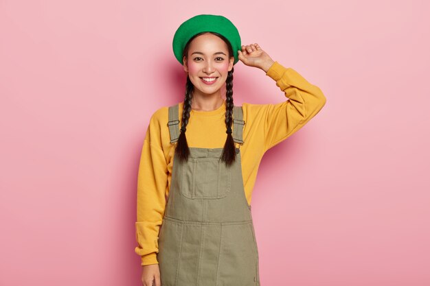 Felice modello femminile asiatico con guance rosse, ha una piacevole espressione del viso, indossa berretto elegante, felpa gialla e sarafan in denim