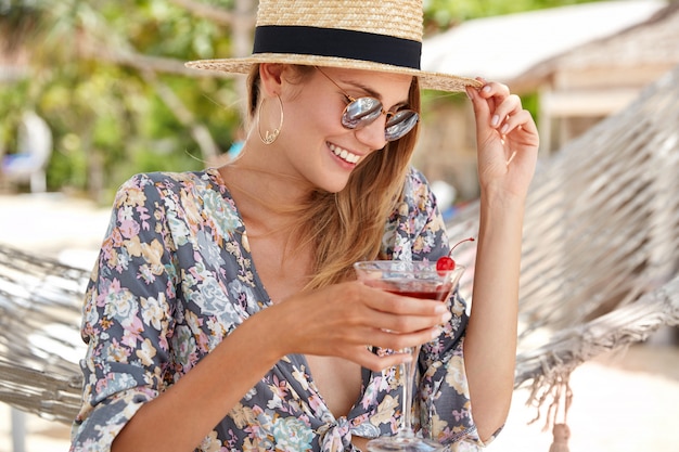 Felice modello femminile rilassato si sente eccitato mentre ricrea all'aperto nel caldo paese tropicale, indossa una camicia stampata a fiori e un cappello di paglia, occhiali da sole alla moda, beve cocktail di ciliegia fresca,