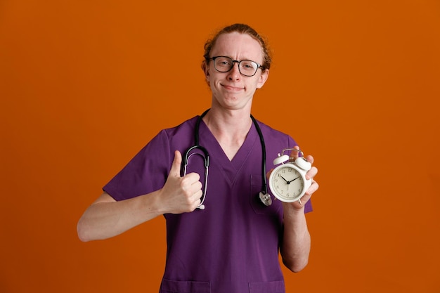 felice mostrando i pollici in su tenendo sveglia giovane medico maschio che indossa uniforme con stetoscopio isolato su sfondo arancione