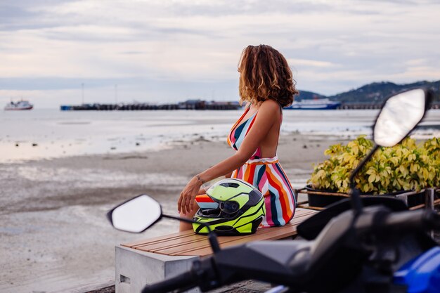 Felice motociclista caucasico donna in abito estivo colorato in vacanza con casco da motociclista