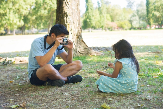 Felice padre asiatico che scatta foto di sua figlia nel parco. Uomo bello che tiene la macchina fotografica della foto che cattura la foto della bambina che si siede sul campo erboso sotto gli alberi. Concetto di svago, hobby e felicità