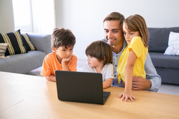 Felice papà seduto al tavolo, abbracciando i bambini e utilizzando il computer portatile. Padre di mezza età caucasico che passa in rassegna in Internet con i bambini adorabili. Infanzia, paternità e concetto di tecnologia digitale