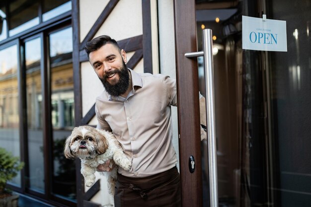 Felice proprietario del bar che tiene un cane mentre apre la porta d'ingresso e guarda la fotocamera