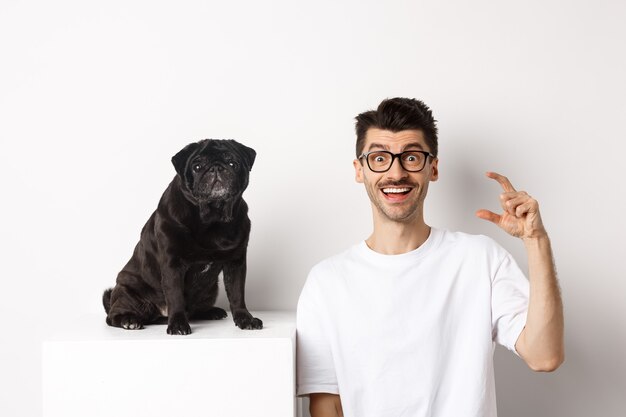 Felice proprietario di un cane seduto vicino a un simpatico carlino nero, sorridente e che mostra una piccola taglia, sfondo bianco.