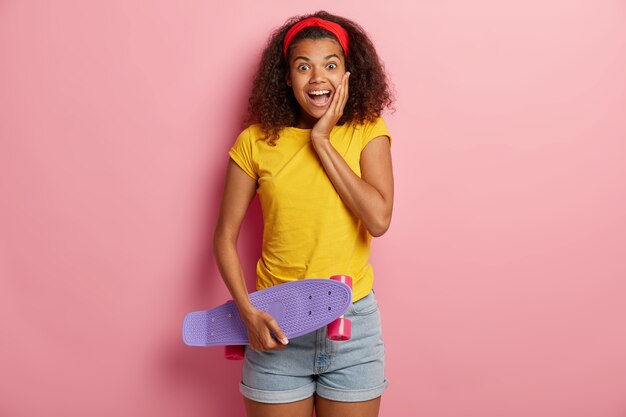 Felice ragazza adolescente con i capelli afro tiene lo skateboard, si diverte con un amico mentre pattina, gode di riposo attivo