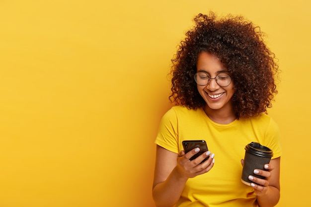 Felice ragazza adolescente con i capelli ricci, tiene in mano un moderno telefono cellulare, caffè da asporto, ordina un taxi tramite applicazione online, digita messaggi di testo, indossa abiti gialli. Persone, stile di vita moderno e tecnologia